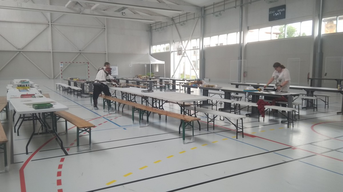 Grandes tables avec des jeux de société dans un gymnase et 2 personnes qui installent des jeux de société