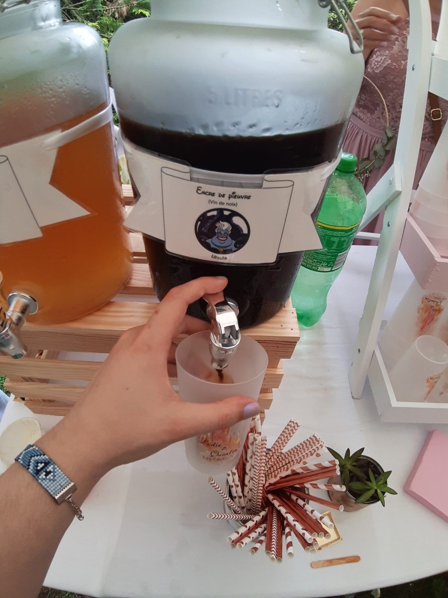 Bonbonne de vin de noix avec une étiquette Ursula de La Petite Sirène, une main actionnant le robinet dans un ecocup à l'effigie du mariage